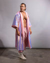 A man modeling a long, iridescent pink and lavender sequined robe with pink trim, paired with pink pants and off-white boots, standing against a plain gray studio backdrop.