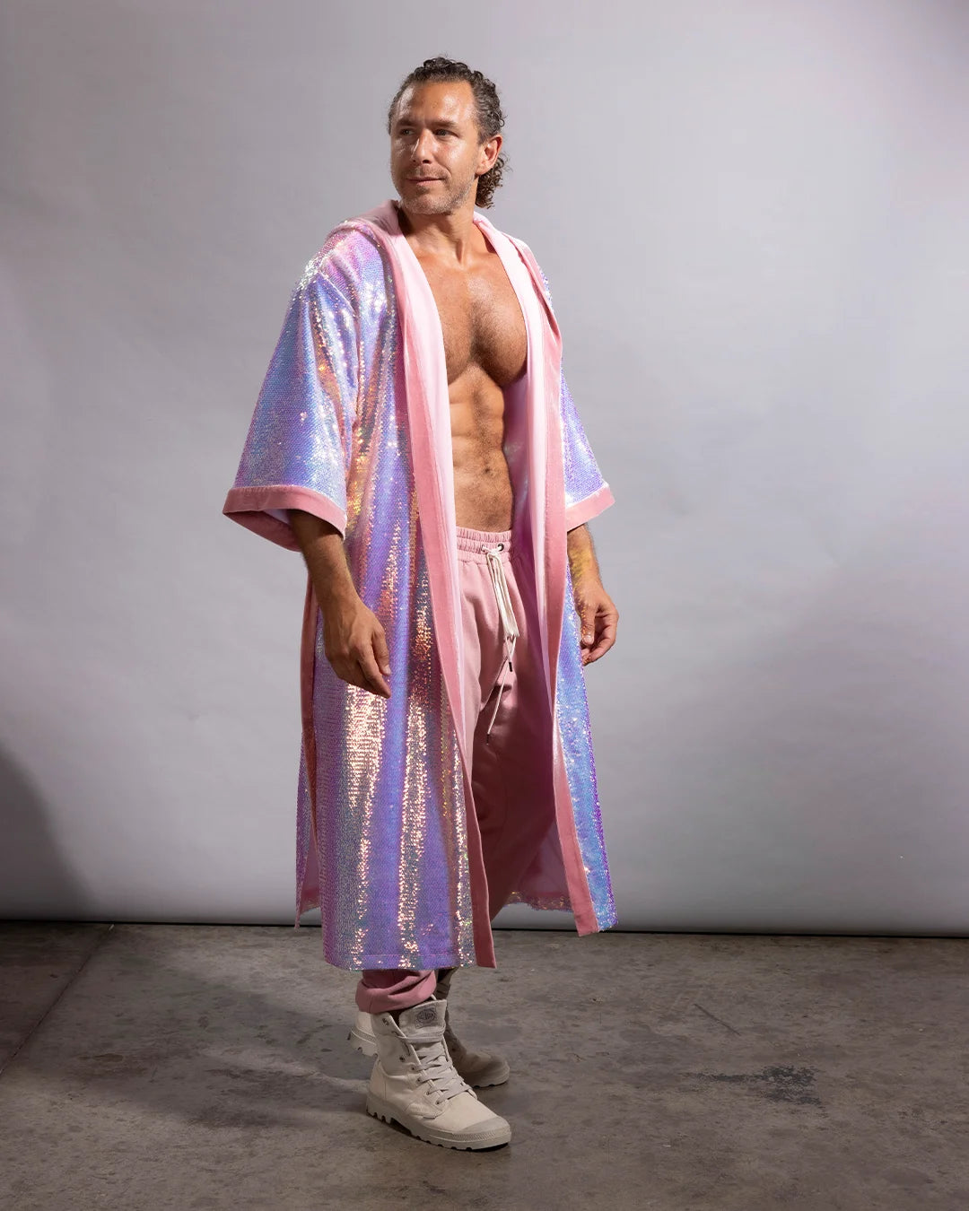 A man modeling a long, iridescent pink and lavender sequined robe with pink trim, paired with pink pants and off-white boots, standing against a plain gray studio backdrop.