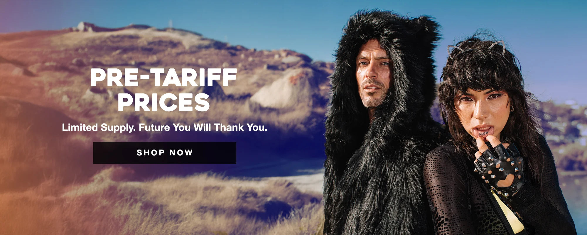 Two models pose outdoors in edgy faux fur fashion against a scenic desert-like background. The man wears a black furry hooded garment with bear-like ears, while the woman wears studded gloves, a sheer black outfit, and sparkly cat ear headband, lifting her lip to reveal a tattoo that says "MEOW." Bold text reads “PRE-TARIFF PRICES – Limited Supply. Future You Will Thank You.” with a “SHOP NOW” button underneath.