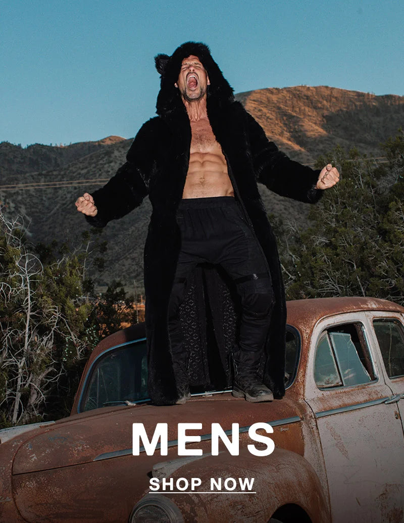 A shirtless man stands triumphantly on the hood of a rusty vintage car in a mountainous desert landscape, wearing a long black faux fur coat with bear ears on the hood. His arms are raised and fists clenched as he yells with intensity. Bold white text at the bottom reads “MENS” with a “SHOP NOW” button underneath.