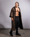 A shirtless man wearing a long, open-front black robe covered in dense bl gold sequins, paired with black pants and black boots, standing against a plain light gray studio backdrop.ck and 