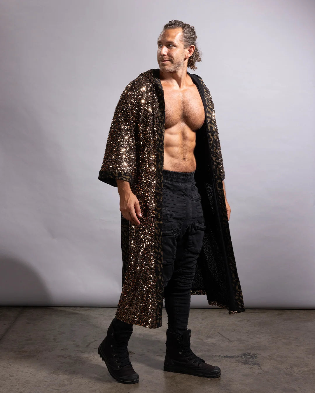 A shirtless man wearing a long, open-front black robe covered in dense bl gold sequins, paired with black pants and black boots, standing against a plain light gray studio backdrop.ck and 