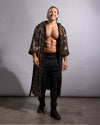 A shirtless man wearing a long, floor-length black sequined robe over black pants and black boots, standing against a plain gray studio background.