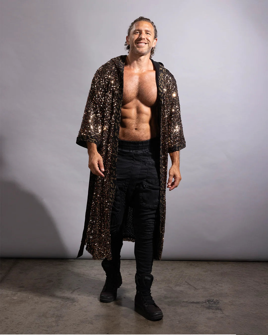 A shirtless man wearing a long, floor-length black sequined robe over black pants and black boots, standing against a plain gray studio background.