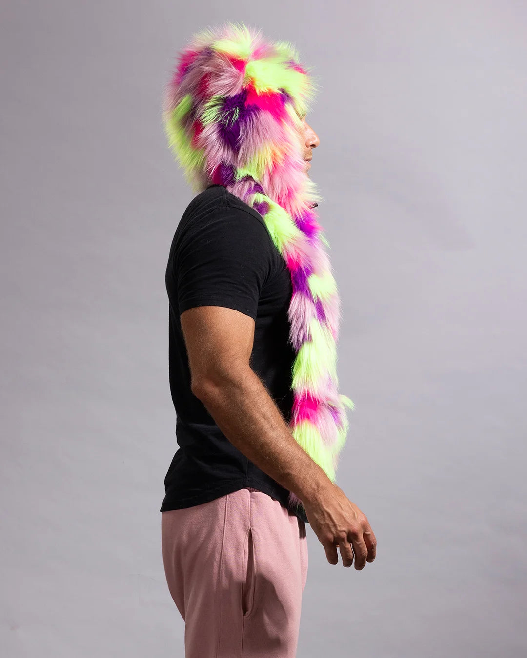 A man in profile wearing a vibrant neon pink, purple, and lime green faux fur hood and long scarf against a plain gray studio background.