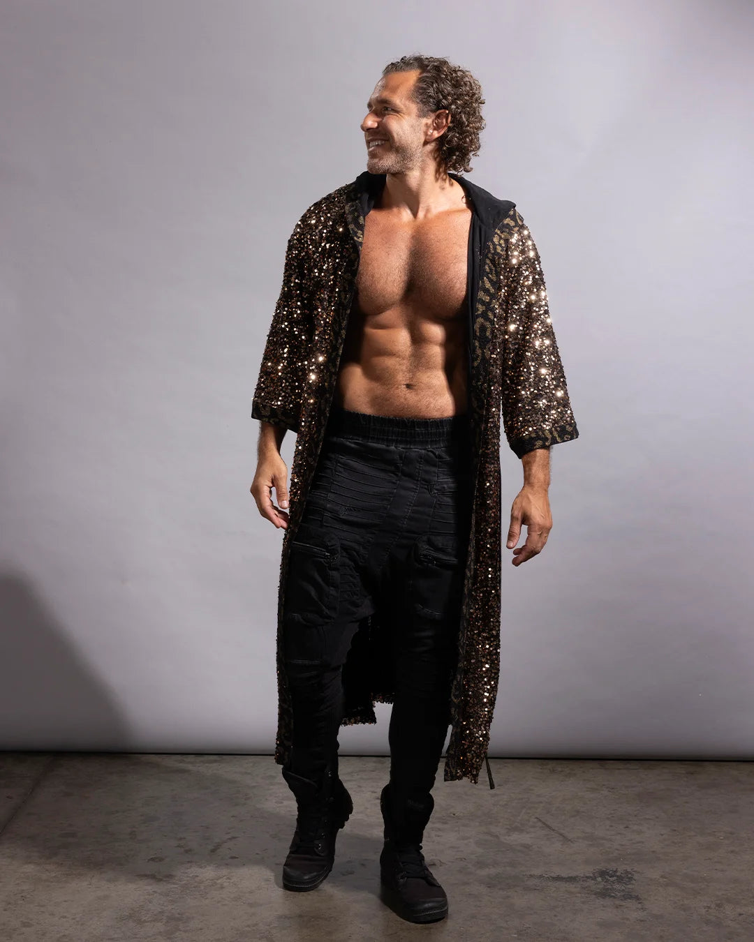 A man modeling a long, open-front black and gold metallic sequined festival kimono robe with three-quarter sleeves, worn over a black shirt and black pants, standing against a plain gray studio backdrop.