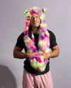 A man wearing a bright neon pink, green, and purple faux fur hood with animal ears and a hand-warmers, over a black t-shirt and pink pants, against a plain gray studio background.
