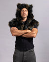 A man wearing a black faux-fur shawl over a black t-shirt and dark gray pants, standing with arms crossed against a light gray studio background.