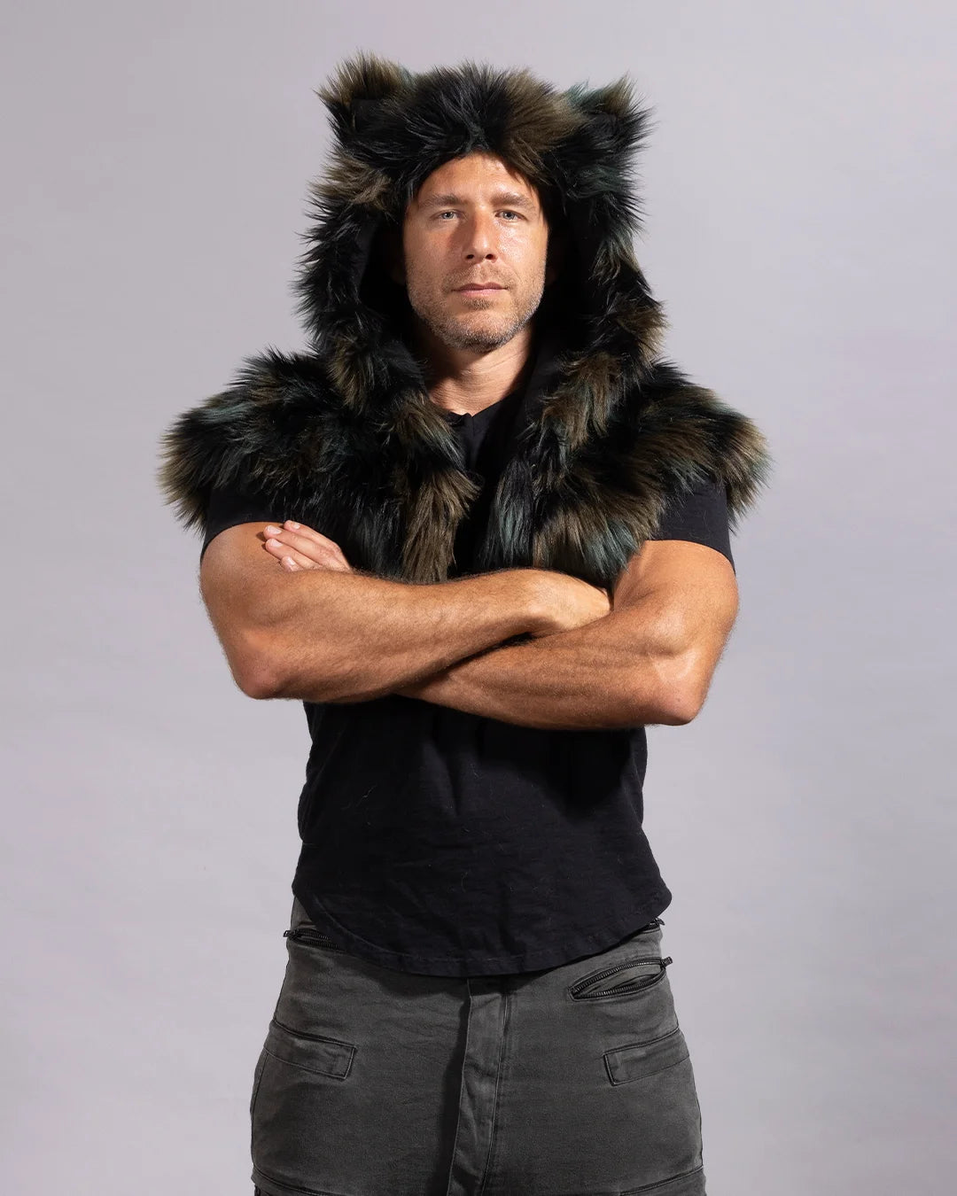 A man wearing a black faux-fur shawl over a black t-shirt and dark gray pants, standing with arms crossed against a light gray studio background.