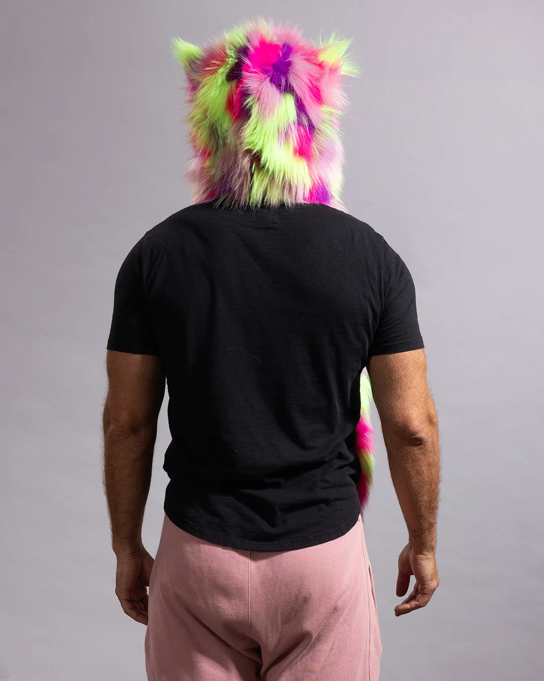 A man viewed from behind wearing a vibrant, fluffy animal-style hood in neon pink, lime green, and purple, paired with a black t-shirt and dusty pink pants, against a solid gray studio background.