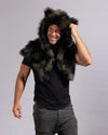 A man wearing a dark brown and black faux fur hooded shawl over a black t-shirt and gray cargo pants, posing against a light gray studio background.