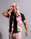 A man wearing a vibrant, multicolored faux fur hooded scarf with pink, green, and purple patches over a black t-shirt and light pink sweatpants, set against a plain gray studio background.