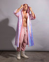 A man modeling a long, iridescent pink kimono coat with a hood, paired with pink sweatpants and off-white boots, standing against a gray studio backdrop.