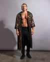 A shirtless man wearing a long, open-front black sequined robe over black pants and black boots, standing against a plain light gray studio background.