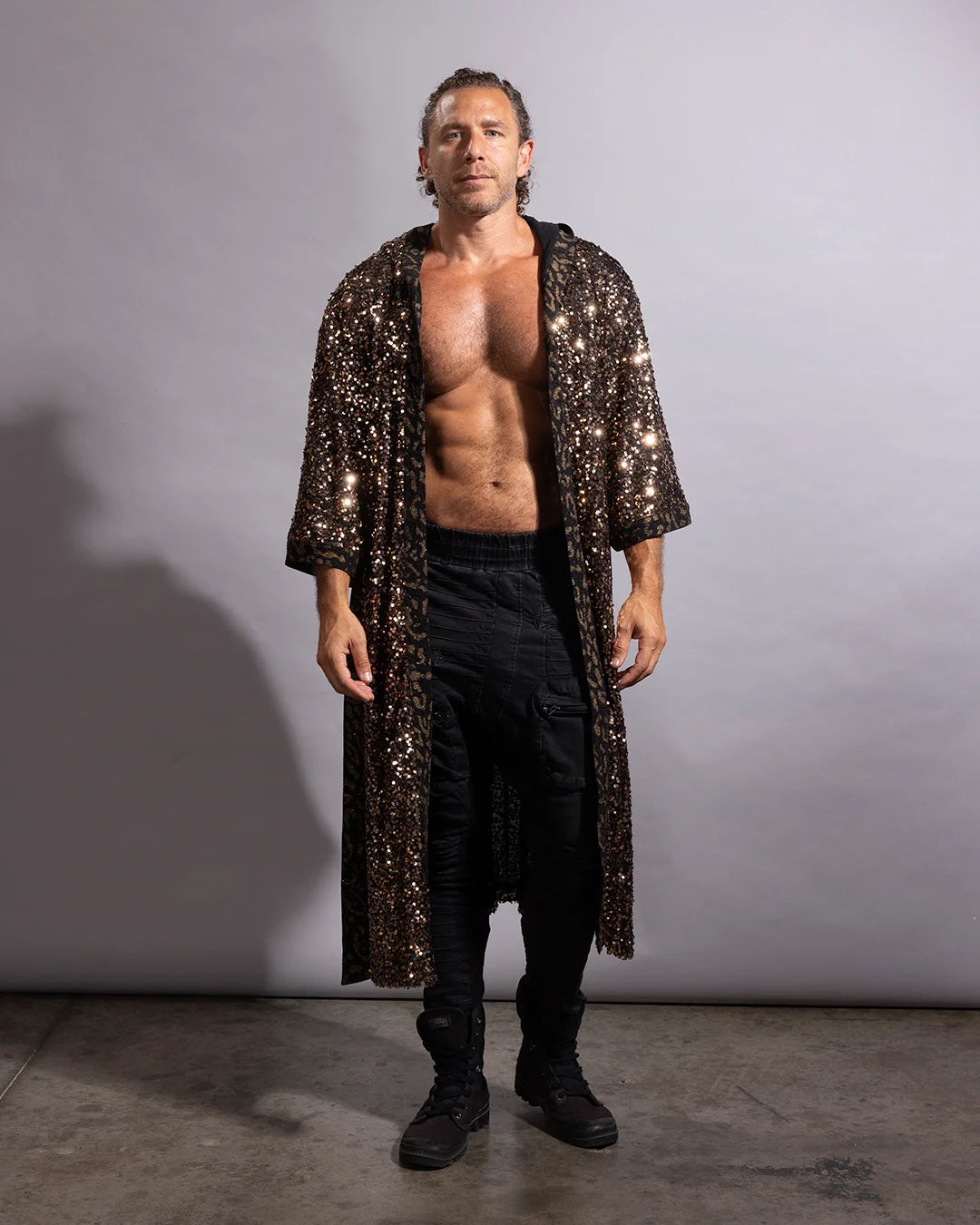 A shirtless man wearing a long, open-front black sequined robe over black pants and black boots, standing against a plain light gray studio background.