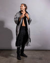 A man stands against a plain gray studio wall wearing a long open-front robe with a silver and black patterned exterior and black lining, paired with black pants and black boots.