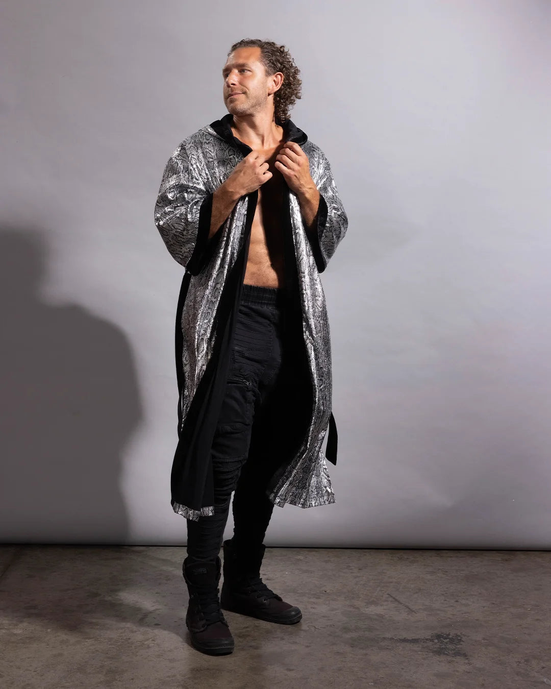 A man stands against a plain gray studio wall wearing a long open-front robe with a silver and black patterned exterior and black lining, paired with black pants and black boots.