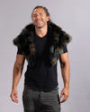 A smiling man wearing a black t-shirt and dark gray cargo pants, styled with a dark brown and black faux fur shawl with teal undertones, against a solid gray studio background.