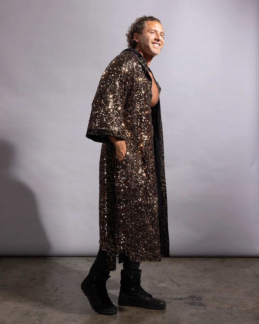 A man wearing a long, black and gold sequined kimono robe with wide sleeves and black boots, standing against a plain gray studio backdrop.