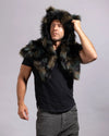 A man wearing a dark brown and black faux fur hooded shawl over a black t-shirt and gray pants, posing against a light gray studio background.