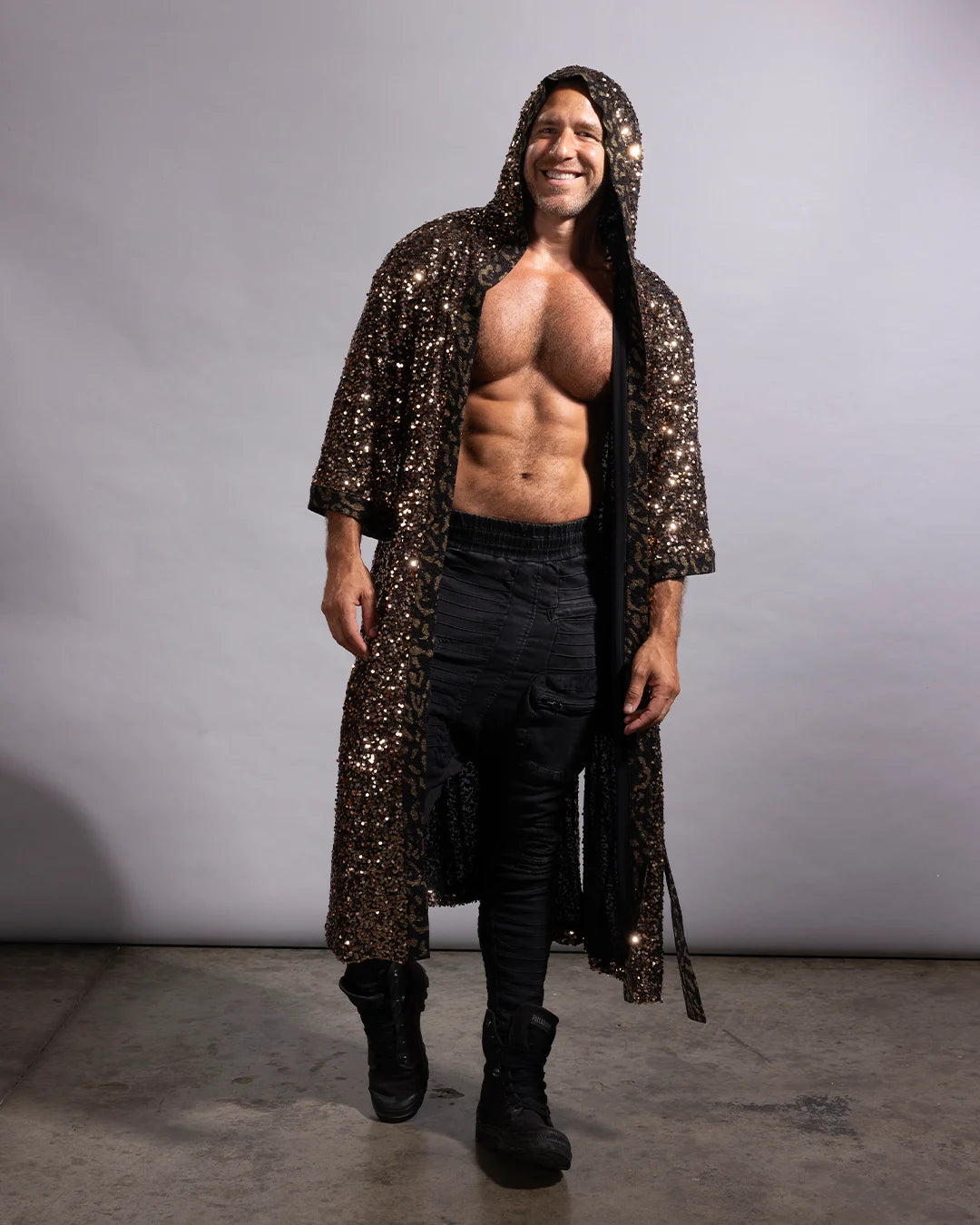 A man wearing a long, hooded, floor-length black sequined robe over a bare torso, paired with black pants and black boots, standing against a plain light gray studio backdrop.