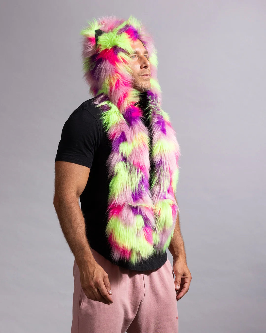 A man wearing a vibrant, multicolored faux fur hood with neon pink, green, and purple tones, styled over a black t-shirt and pink sweatpants against a plain gray studio background.
