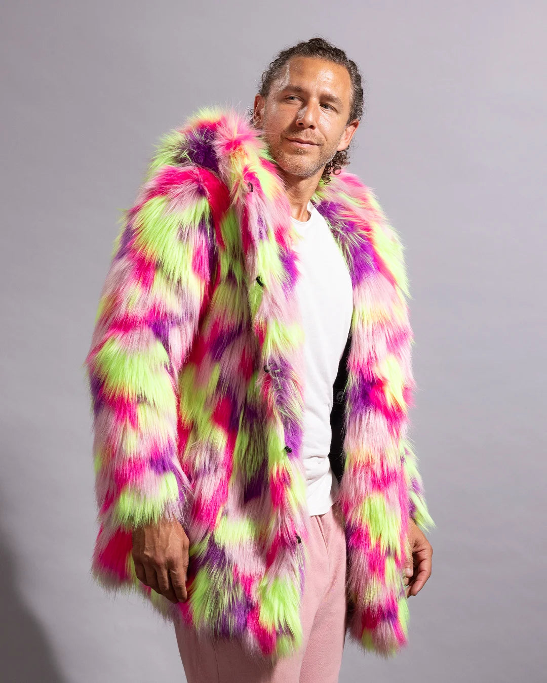 A man wearing a vibrant, multicolored faux fur coat with pink, green, and purple patterns, styled over a white t-shirt and light pink trousers, standing against a plain gray studio background.