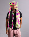 A man wearing a neon multicolor faux fur hooded scarf with black paw-print accents, a black t-shirt, and pink sweatpants, photographed against a solid gray studio background.
